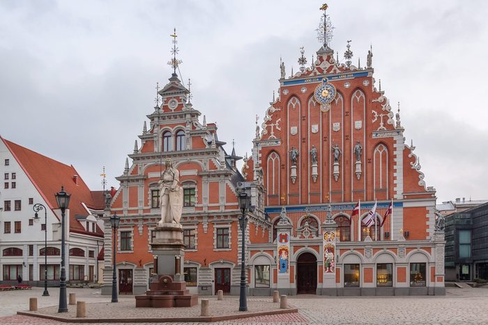 Statue stands in front of ornate red-brick building adorned with flags and decorative statues; cobblestone square surrounds structure under overcast sky.
