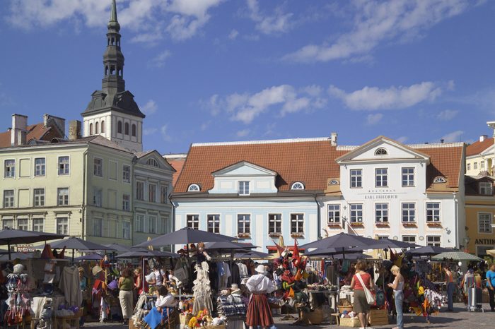 Market stalls display crafts, bustling with people, set against colorful historic buildings under a clear blue sky.