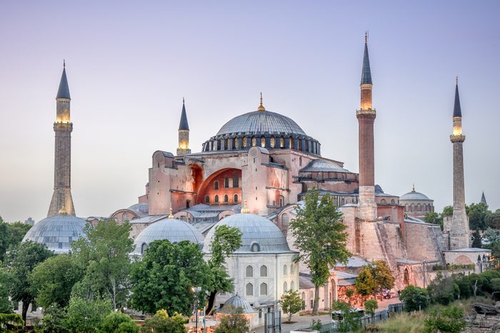 Historic domed building stands, surrounded by tall minarets, against a twilight sky, with trees and smaller structures in the foreground.