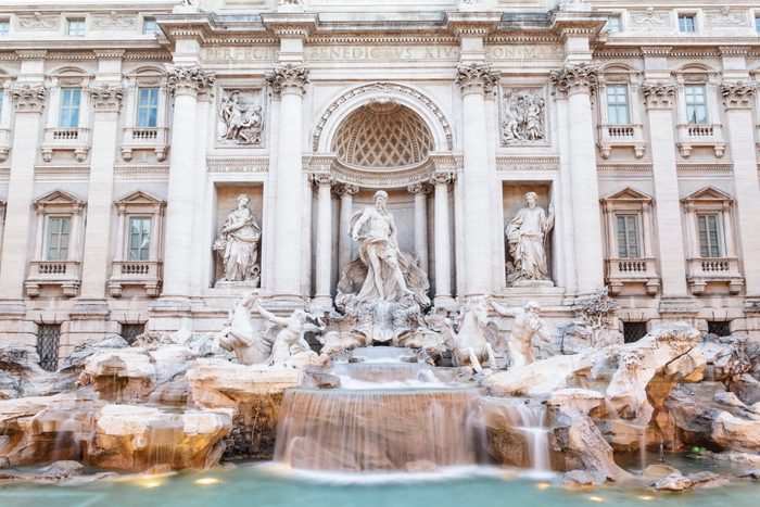 Statue stands atop cascading fountain; intricate carvings adorn building facade in classical style. Latin text inscribed above.