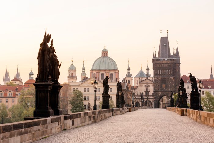 Statues stand still on an old cobblestone bridge, surrounded by historic buildings with domes and spires under a softly lit sky.
