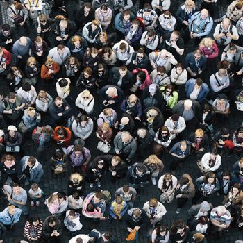 People stand closely together, some looking at devices, in a crowded cobblestone area, suggesting an event or gathering.