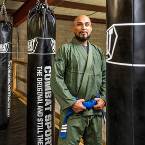 A person stands confidently wearing a green martial arts uniform, holding a blue belt, surrounded by black punching bags in a gym setting.