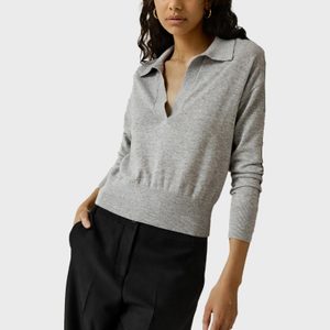 Person wears gray sweater and black pants, standing against a plain backdrop with hand in pocket.
