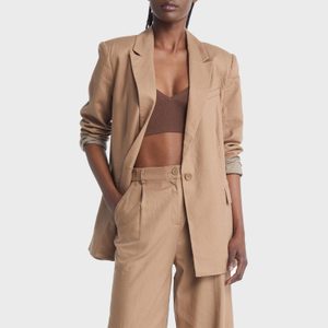 Person wears a tan suit with rolled sleeves, resting their hand in a pocket, standing against a plain white background.