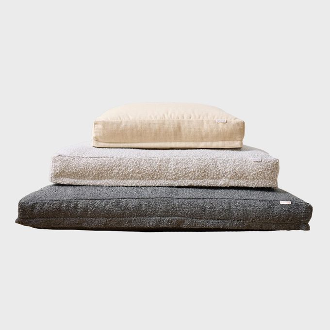 Three stacked cushions in beige, light grey, and dark grey. The cushions are textured, positioned on each other, against a plain background.