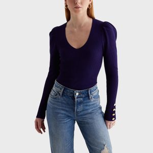 A person wearing a deep purple sweater with gold button cuffs stands, paired with blue jeans, against a plain background.