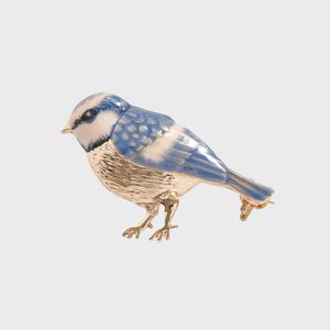 A blue and gold bird pin rests on a plain light gray surface, displaying intricate feather patterns and metallic highlights.