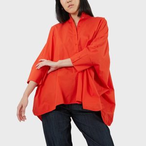 A person wearing an oversized, bright red shirt poses with arms gracefully crossed, set against a plain white background.