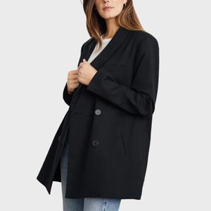 Woman wearing black blazer over white shirt, adjusting it, with light blue jeans against plain gray background.