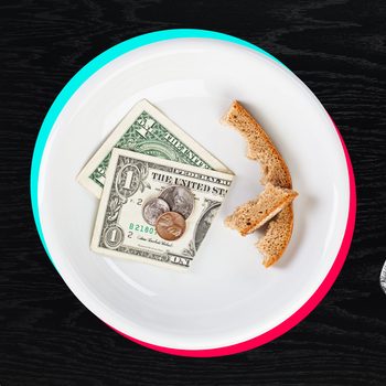 A plate holds U.S. dollar bills, coins, and bread crusts, surrounded by a fork and knife on a dark wooden table.