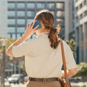 Person holding a phone to their ear, standing outdoors in a city setting, surrounded by buildings and trees.