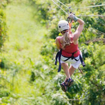 Person zip-lining through lush, green forest; wearing a helmet, harness, and red top.