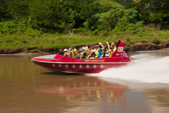 Red speedboat racing on a river, with passengers wearing life jackets, surrounded by lush green trees and vegetation. Text: 