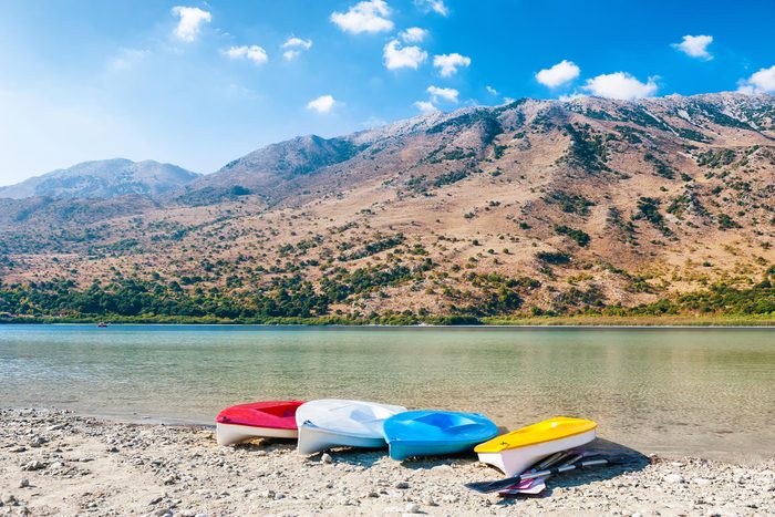 Four colorful kayaks rest on a rocky lakeshore, surrounded by clear water and a mountainous landscape under a bright blue sky.