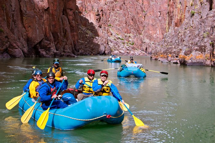 Blue rafts carry people paddling through a river, surrounded by steep canyon walls. Everyone wears helmets and life jackets.