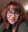 A person with glasses smiling, framed by medium-length brown hair, indoors in softly lit surroundings.