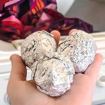 Hand holds crumpled aluminum foil balls near a colorful fabric, likely in a laundry setting, suggesting a potential use in the washing process.