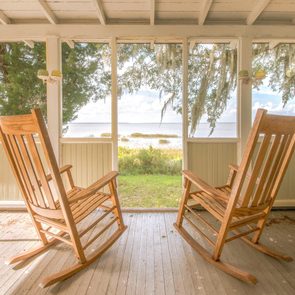Two wooden rocking chairs face each other on a porch, overlooking a serene lakeside view with trees and moss hanging nearby.