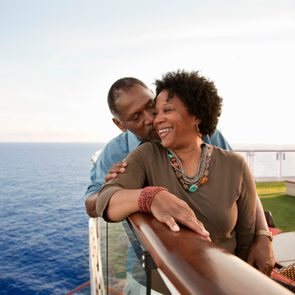 A couple embraces on a cruise ship deck, enjoying ocean views, with another person seated in the background.