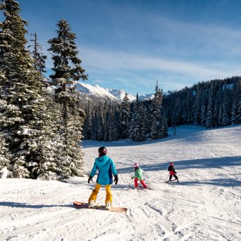 Snowboarder descends slope, flanked by two skiers, surrounded by snow-covered pine trees under a clear blue sky.