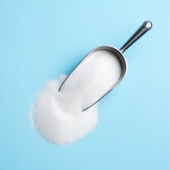 A metal scoop pours white sugar onto a light blue surface.