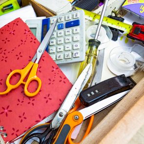 Scissors rest on a red notebook amidst clutter, including a calculator, screwdriver, measuring tape, staples, and tape in a messy drawer.