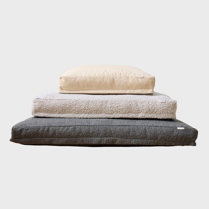 Three stacked cushions of varying sizes. The top is beige, middle is light gray, and bottom is dark gray. Simple background.