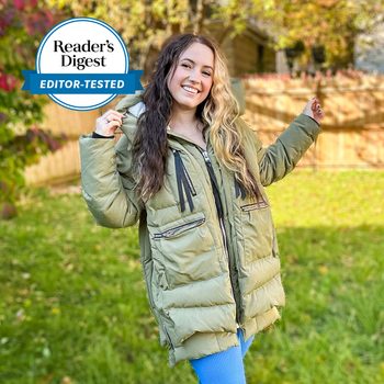 Person wearing a green puffer coat smiles in a garden with trees and wooden fence. Text reads "Reader's Digest Editor-Tested."