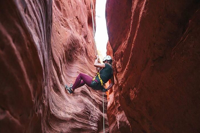 Person rappelling down a narrow, red rock canyon, wearing a helmet and climbing gear, surrounded by steep, rugged sandstone walls.