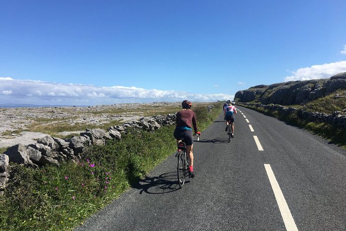 Cyclists ride along a winding road bordered by stone walls and wildflowers under a clear blue sky.