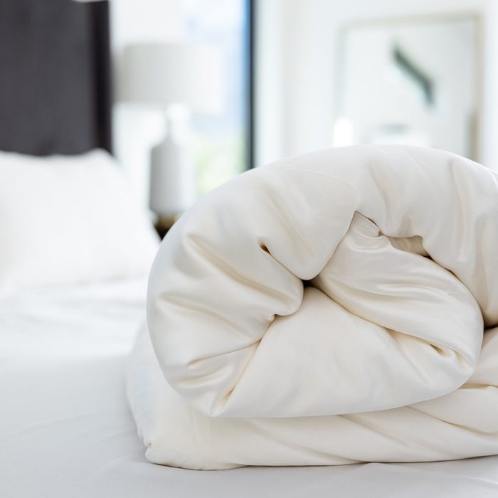 A white blanket is neatly folded on a bed, with a blurred bedroom background featuring a lamp and a window.