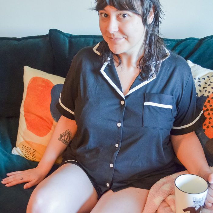 Person in black pajamas sits on a couch, holding a mug. Bright pillows are in the background, adding color to the scene.