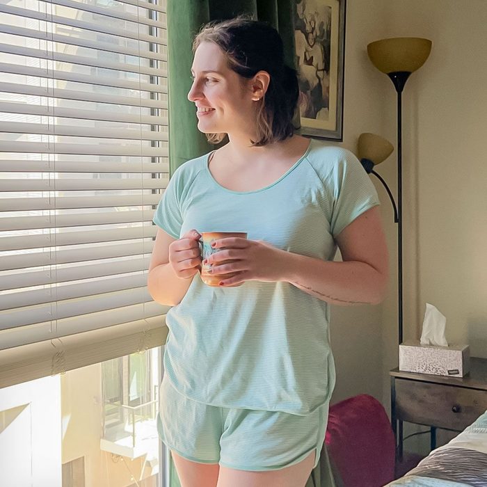 A person holding a mug, gazing out a sunlit window in a cozy room with a floor lamp and tissue box nearby.