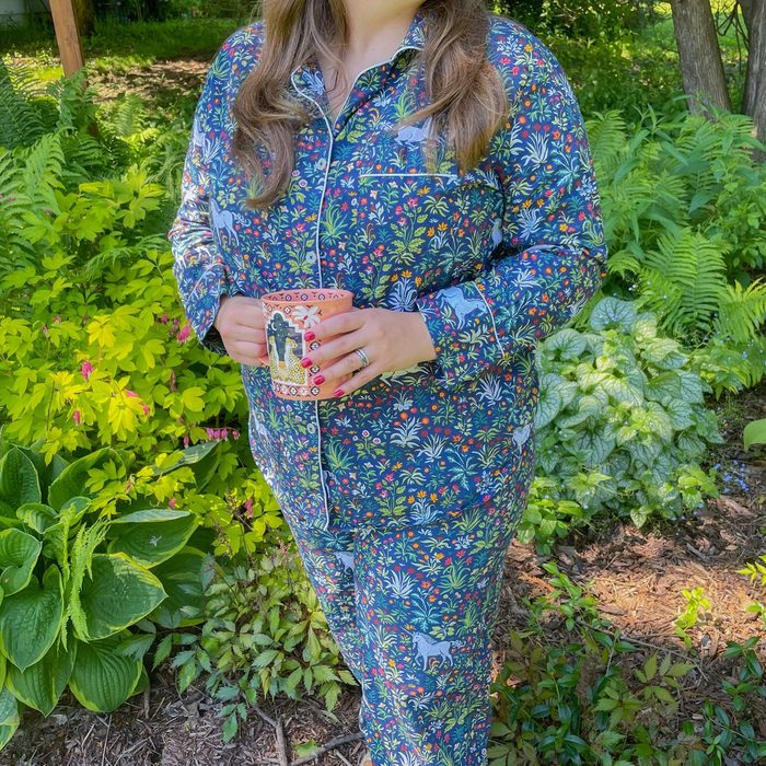 Person wearing colorful pajamas holds a mug, standing in a lush garden with various green plants and ferns around.