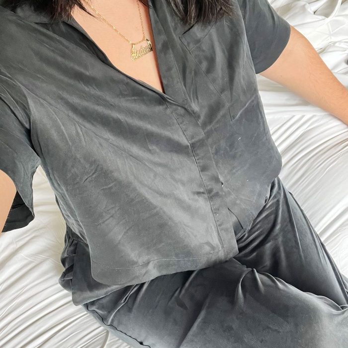 Person wearing dark silk pajamas sits on a white bed, showing a gold necklace with a name.
