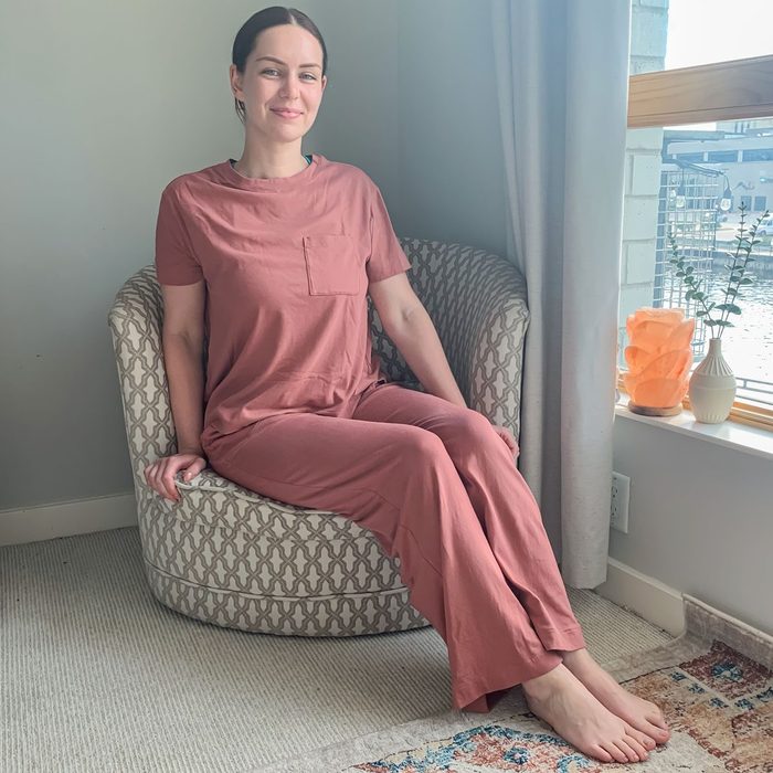 A person in pink loungewear sits on a patterned chair, near a window with vases on the sill, in a softly lit room.