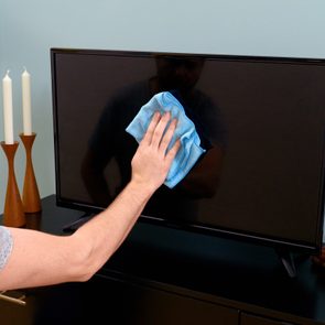 A person cleans a television screen with a blue cloth in a room with candles, a lamp, and a plant on a shelf.