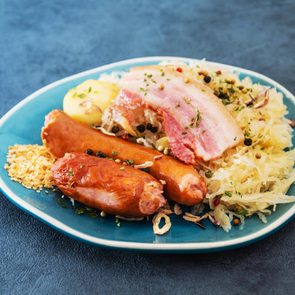 A plate holds sausages, sauerkraut, bacon, and potato garnished with herbs, on a textured blue surface.