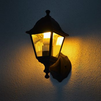 Wall-mounted lantern emits warm light, illuminating a textured, dark wall in a dimly lit setting.