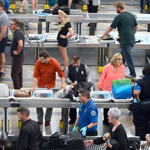 Travelers place items in trays at a bustling airport security checkpoint. Security officers inspect bags, while passengers wait and prepare belongings on conveyor belts.