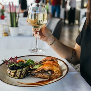 Plate holds grilled prawns and vegetables; person lifts a wine glass in an elegant dining setting.