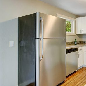 Refrigerator stands closed in a kitchen corner, near a window and white cabinets, with a wooden floor and beige walls.