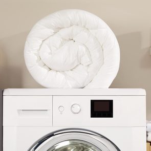 A rolled-up white comforter rests atop a front-loading washing machine; towels and bottles are neatly arranged on surrounding shelves.