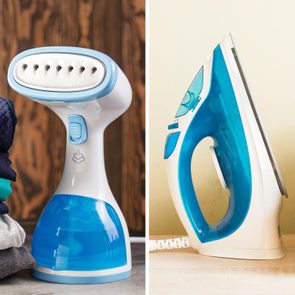 A handheld steamer releases steam next to folded clothes; an upright electric iron sits on a wooden surface adjacent to a light-colored wall.