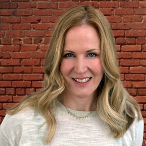 A person with long blonde hair smiles in front of a red brick wall, wearing a light gray sweater.