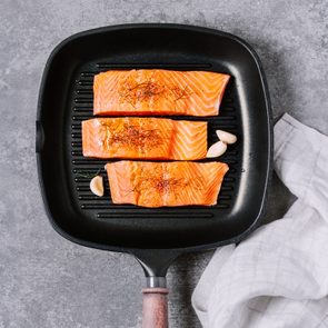 Salmon fillets resting in a black grill pan, garnished with herbs alongside garlic, set against a gray textured surface with a white cloth.