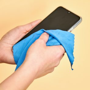 Hands clean a smartphone screen with a blue cloth against a plain beige background.