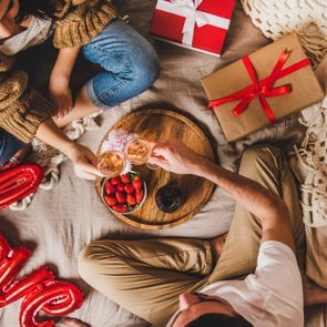 Two people clink glasses over strawberries on a wooden tray, surrounded by pillows, gifts, and a red 