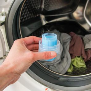 Hand holds blue detergent pod near open washing machine filled with clothes.
